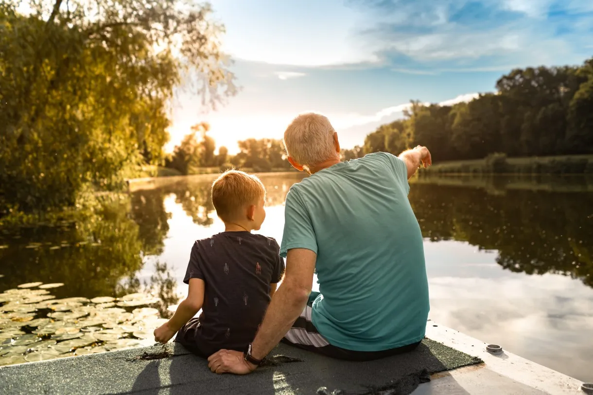 Grandfather and grandson sitting together outdoors on a dock by a lake bonding enjoying nature sunset
